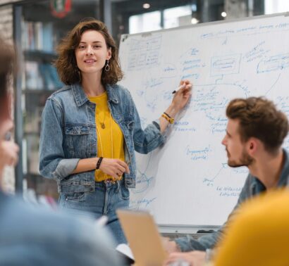 2ï¸âƒ£ A female manager is presenting a marketing plan on a whiteboard during a meeting in the office. A woman wearing a denim jacket and a yellow shirt is standing at the front of the conference room, pointing to a flip chart with a flowchart diagram while talking about a sales strategy to the team members. --ar 3:2 --v 7 Job ID: 74070ca9-c3b1-41cb-a5dd-8c17a695ce36